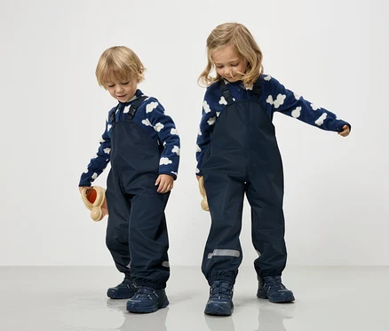 Deux enfants en salopettes bleu marine et pulls nuages bleu et blanc se tiennent sur un fond blanc.