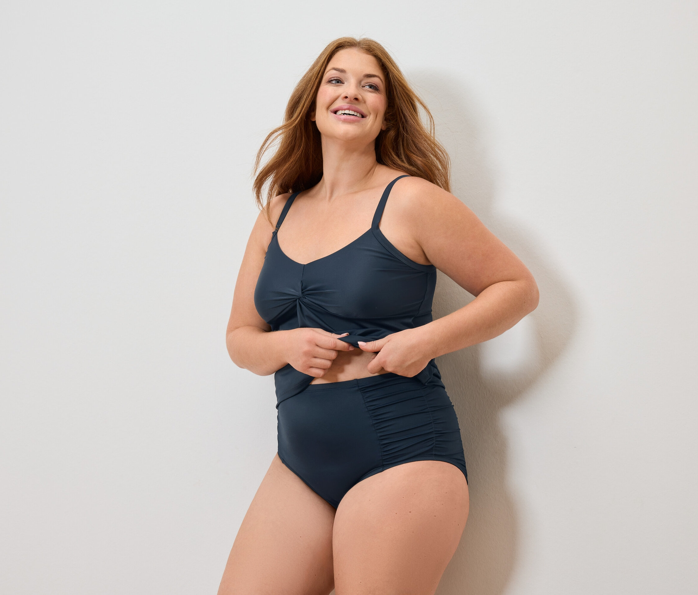 Femme aux longs cheveux roux portant un maillot de bain bleu marine et souriant.