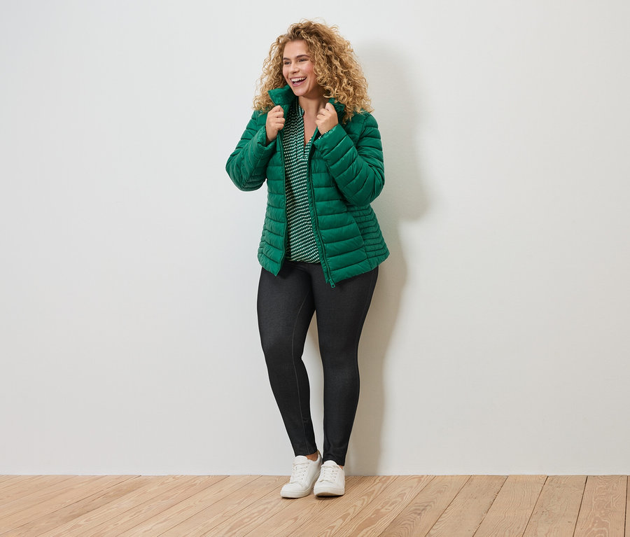 Femme souriante aux cheveux bouclés portant une veste matelassée verte, un chemisier rayé, un legging foncé et des baskets blanches.