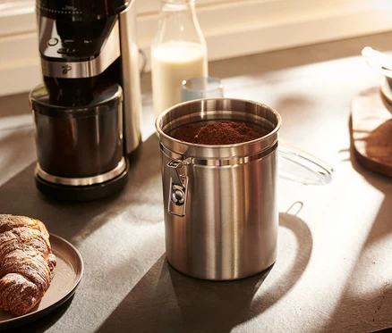 Une boîte à arômes en acier inoxydable est posée sur un comptoir de cuisine à côté d'une machine à café et d'une assiette avec un croissant.