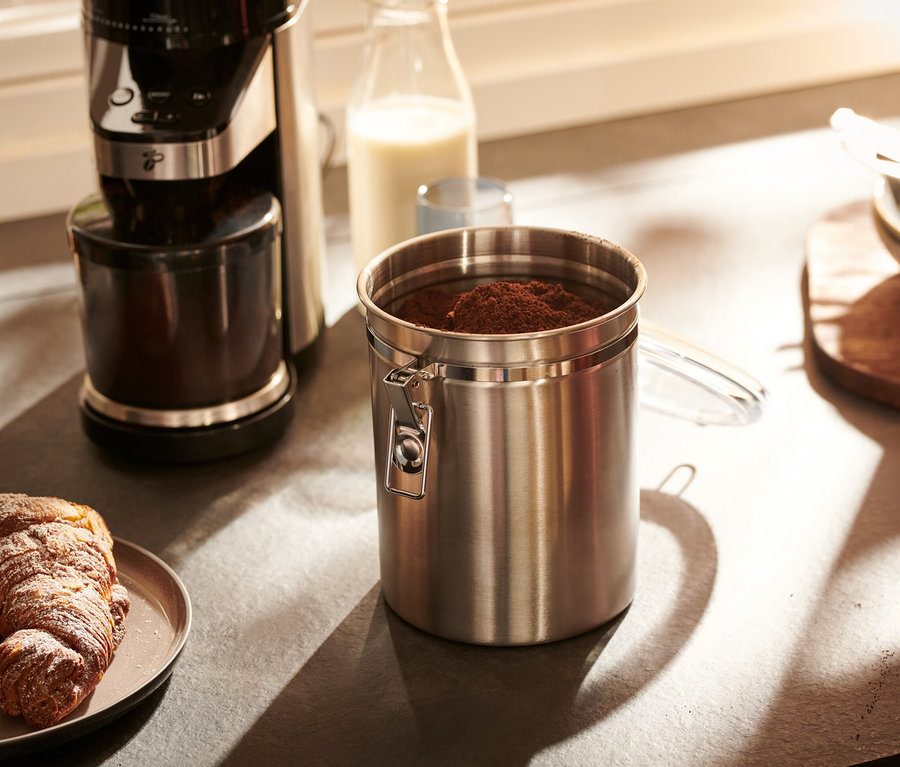 Une boîte à arômes en acier inoxydable est posée sur un comptoir de cuisine à côté d'une machine à café et d'une assiette avec un croissant.