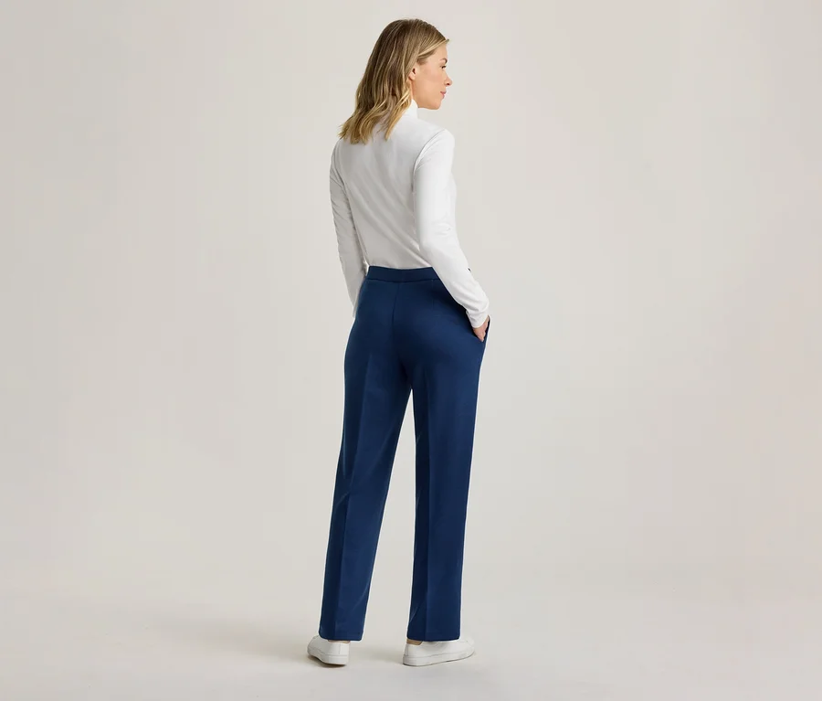 Femme debout de dos, vêtue d'un haut blanc à manches longues et d'un pantalon bleu, les mains dans les poches, regardant vers la droite.