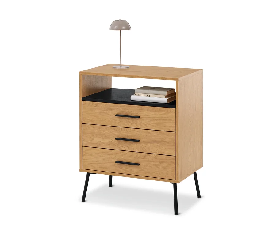 Table de chevet en bois clair avec trois tiroirs et une étagère ouverte, surmontée d'une lampe grise et de livres.