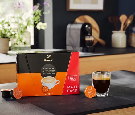 Un paquet de Cafissimo Caffè Crema vollmundig 96 capsules à côté d'un verre de café. À l'arrière-plan, une machine à café Cafissimo pure+, black.