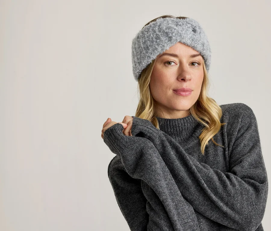 Une femme blonde porte un bandeau en tricot gris et un pull gris, regardant devant elle avec les mains jointes.