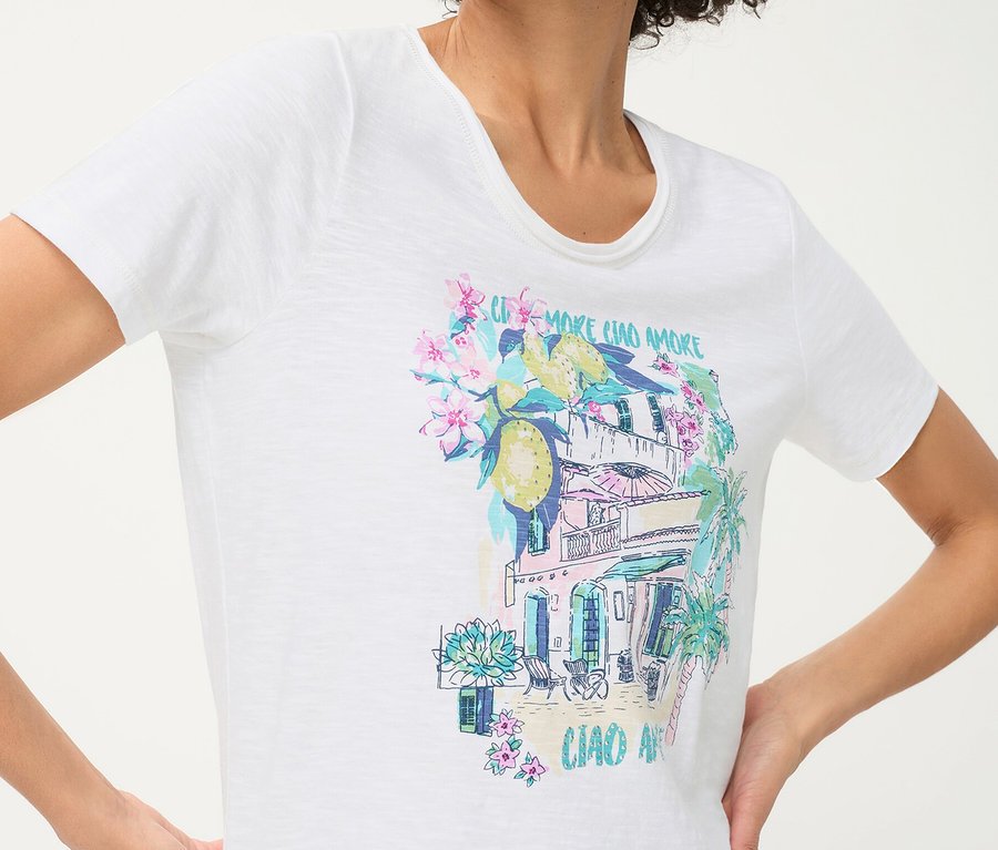 Personne portant un t-shirt Olsen blanc avec un imprimé placé coloré représentant un bâtiment méditerranéen, des citrons, des fleurs et des palmiers.