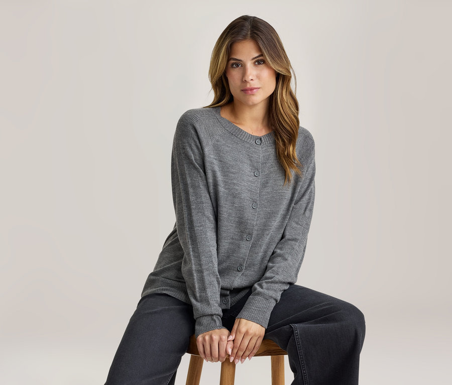 Femme assise sur un tabouret en bois, portant un cardigan gris boutonné et un pantalon large gris foncé, regardant devant elle.