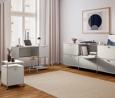 Un bureau à domicile moderne avec des meubles modulaires gris clair. Un bureau avec un ordinateur portable et une lampe noire est placé devant une grande fenêtre. Sous le bureau se trouve un Tabouret avec assise en bois. À gauche, un meuble mobile avec une plante et une bougie. À droite, une longue unité de rangement avec un compartiment ouvert, des livres, un tourne-disque et une œuvre d'art. Un grand tapis recouvre le parquet.