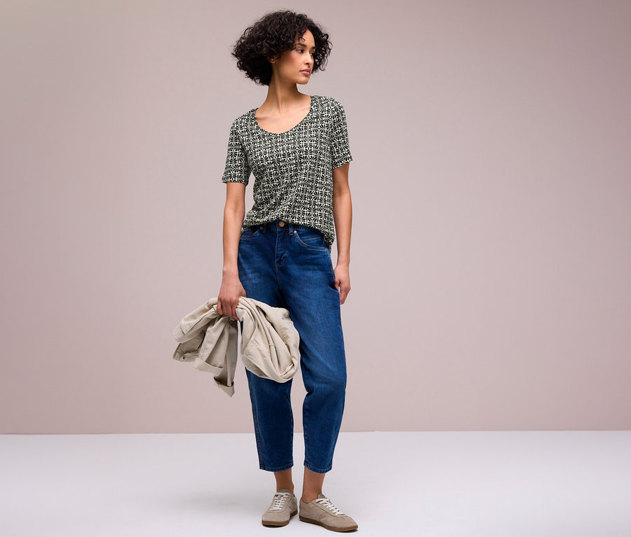 Une femme aux cheveux courts et bouclés porte un T-shirt Street One à motifs noirs et blancs, un jean bleu et des baskets claires. Elle tient une veste beige clair dans sa main gauche et regarde vers la droite.