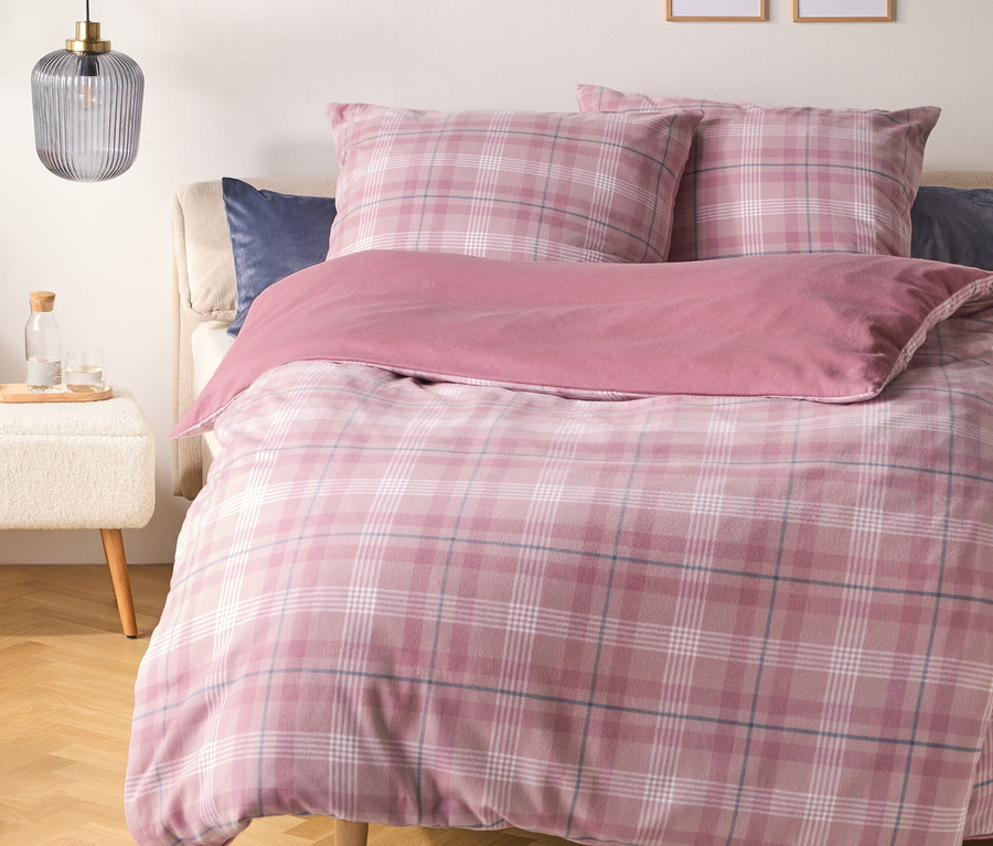 Un lit avec une housse de couette et des oreillers à carreaux roses, et une couverture rose unie. Un oreiller bleu foncé est derrière les oreillers à carreaux. À gauche, un tabouret blanc supporte une bouteille en verre et deux verres, sous une suspension grise nervurée. Deux impressions d'art linéaire encadrées sont accrochées au mur au-dessus du lit.