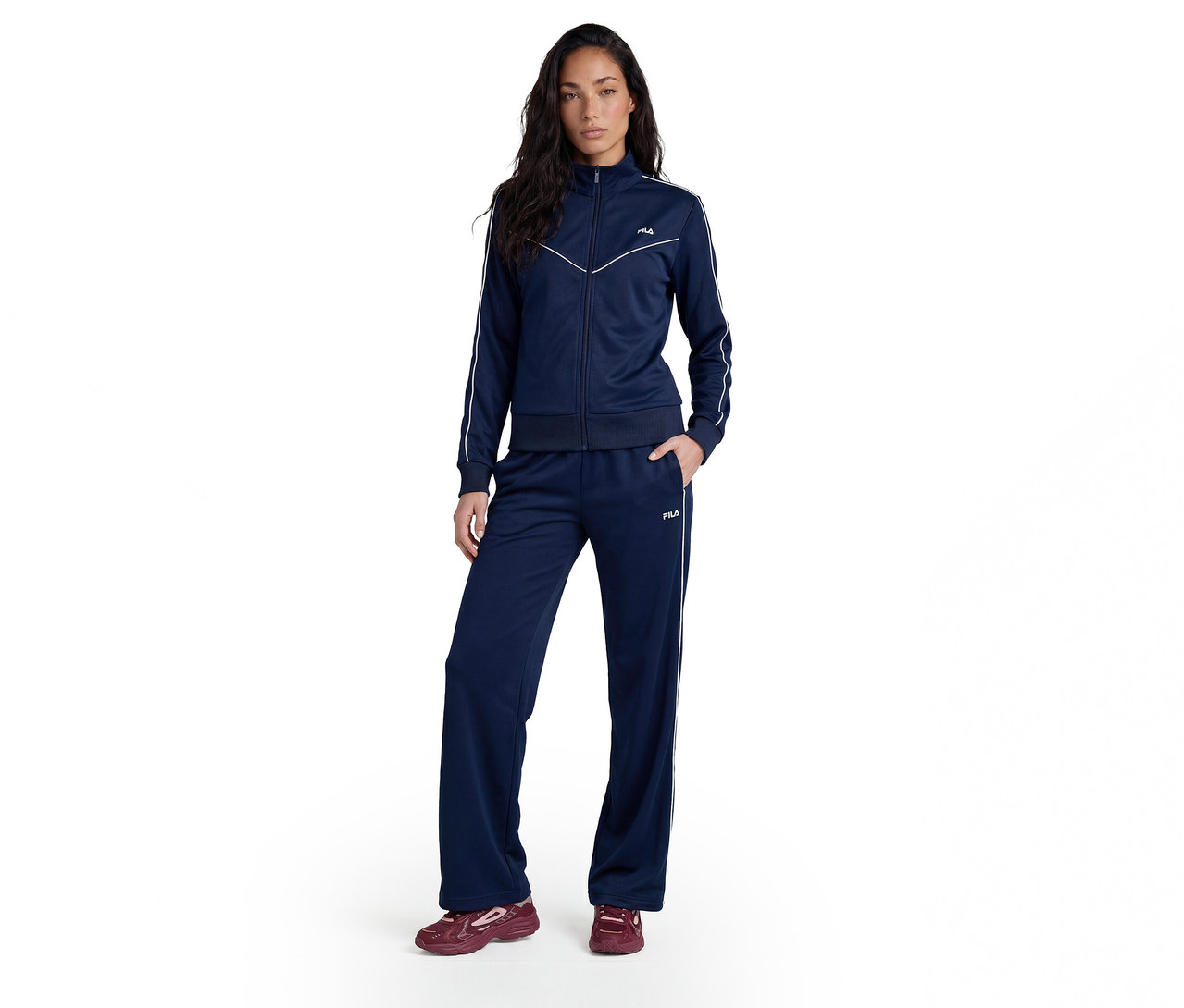Femme debout, vêtue d'un survêtement bleu marine avec des rayures blanches sur les manches et les jambes, et des baskets rouges.
