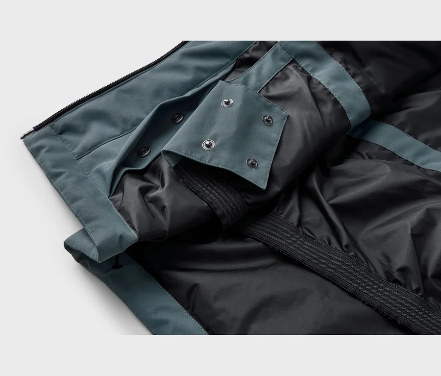 Gros plan d'une veste montrant son tissu extérieur sarcelle et sa doublure intérieure noire. Une fermeture éclair est visible en haut, et un rabat avec quatre boutons-pression est ouvert, révélant la doublure noire et une bande élastique.