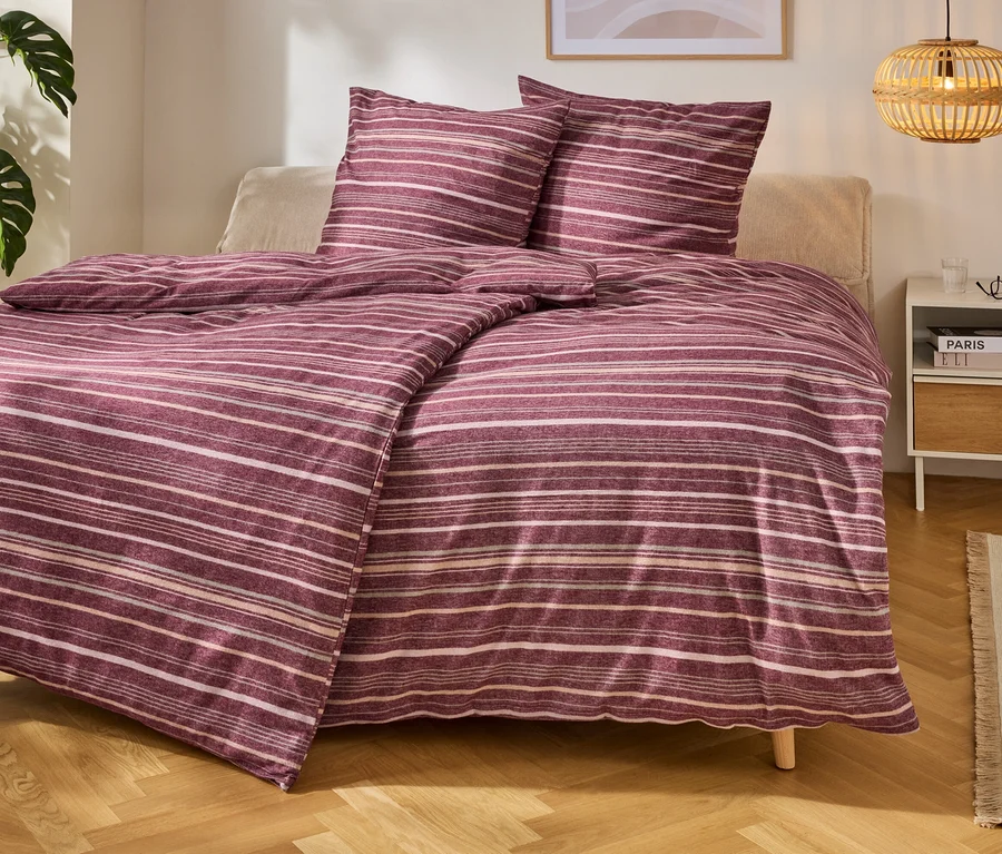 Un lit avec une parure de lit rayée violette et blanche, à côté une table de chevet avec des livres et une lampe. Une plante est à gauche.