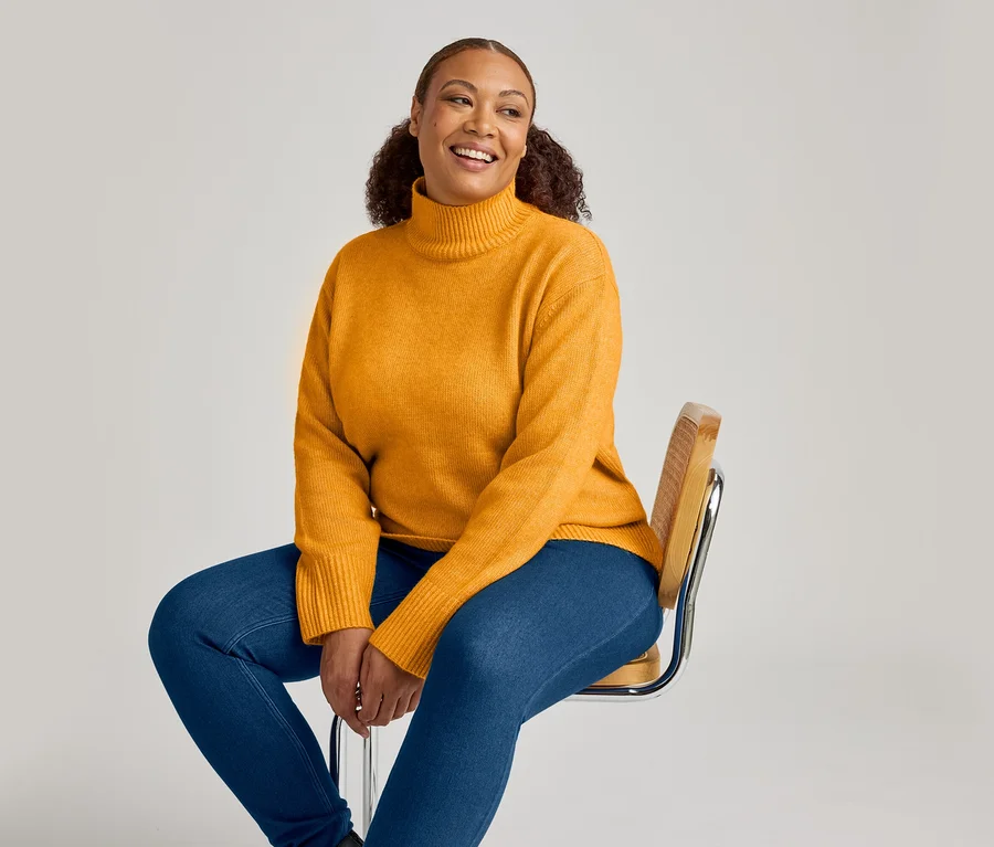 Une femme souriante aux cheveux bouclés est assise sur une chaise, vêtue d'un pull à col roulé jaune moutarde et d'un jegging en denim bleu moyen.