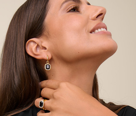 Une femme sourit et touche son cou, elle porte une chaîne Royal Black, des créoles Royal Black et une bague Royal Black.
