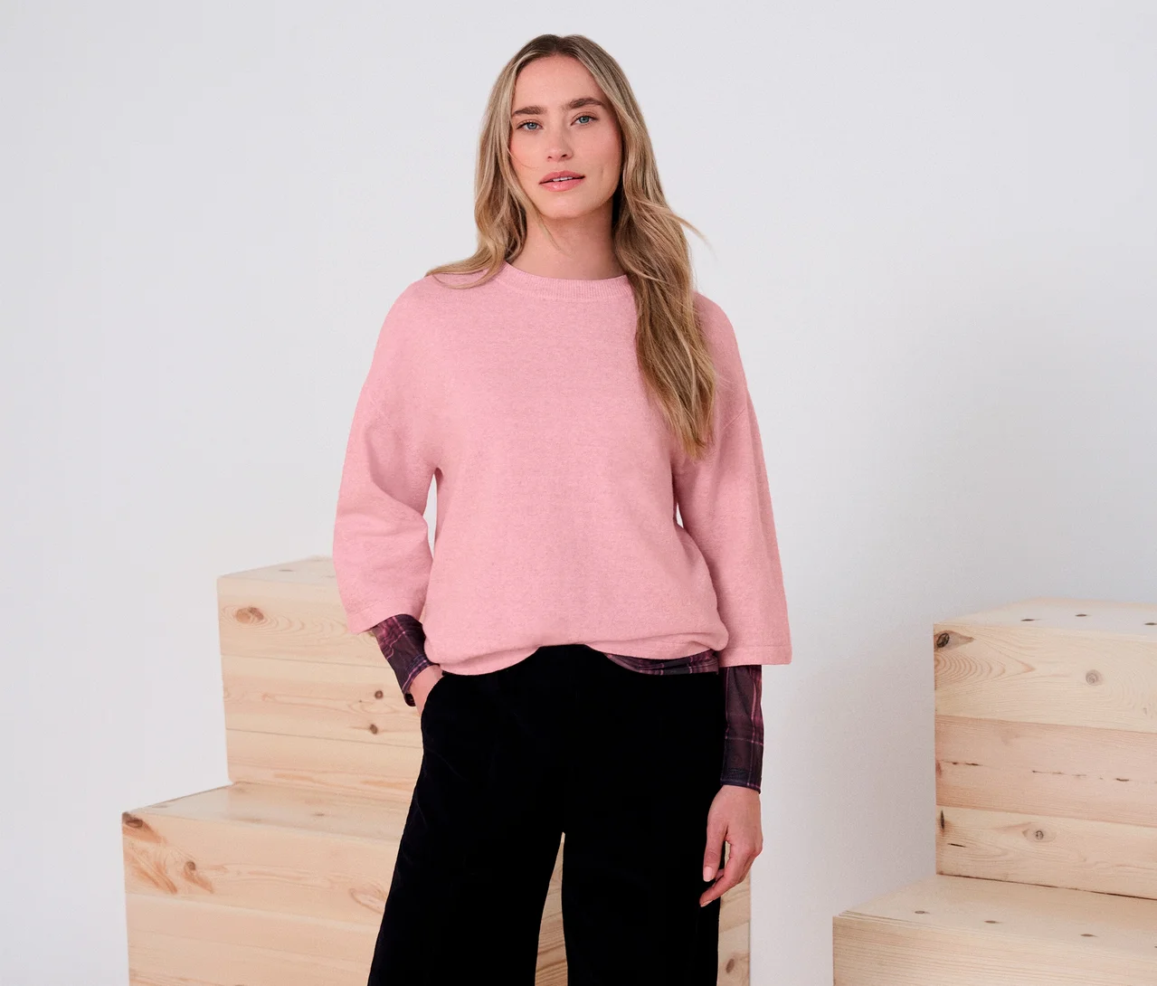 Une femme porte un pull en maille 3/4 Street One rose, une chemise à carreaux et un pantalon noir. Elle se tient devant un fond blanc avec des caisses en bois.