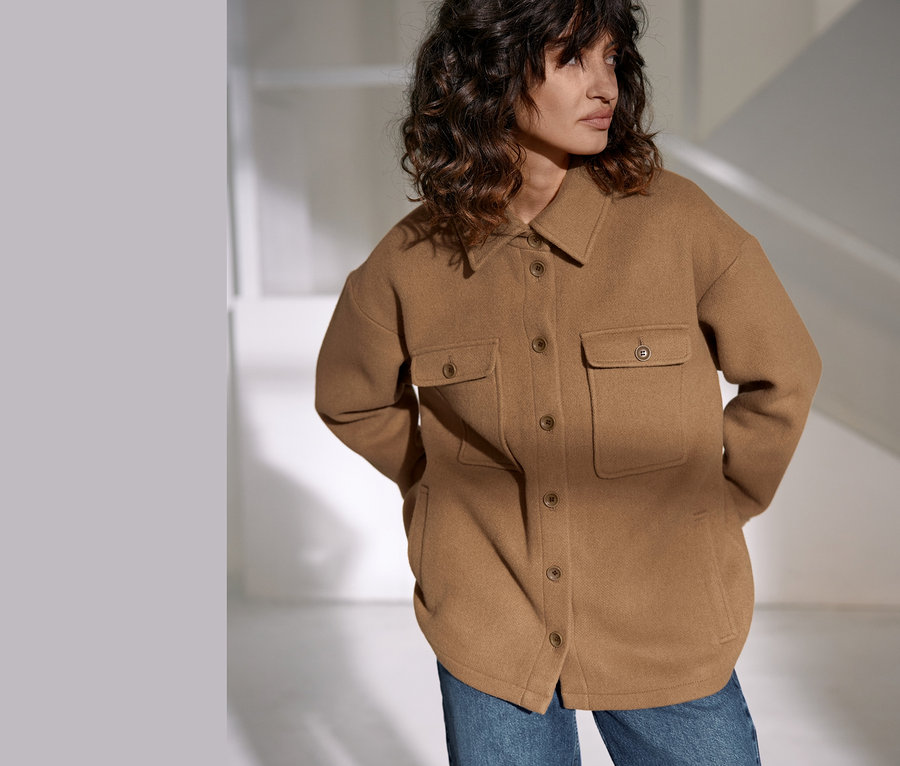 Une femme porte une veste NAH/STUDIO en laine recyclée de couleur camel et un jean.