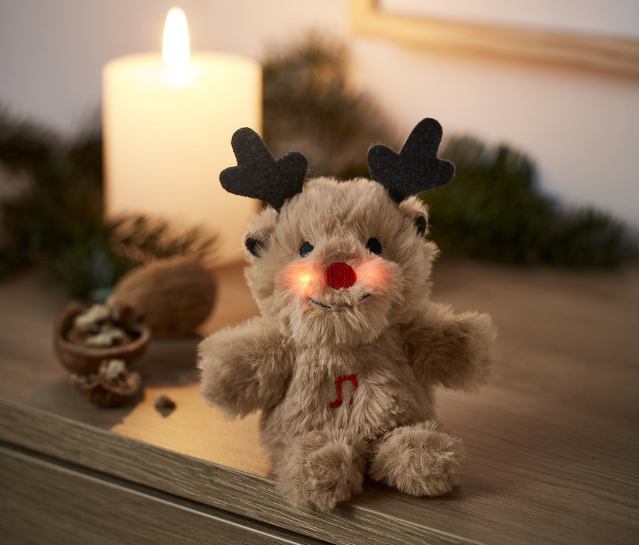 Une mini peluche chantante renne avec un nez rouge est assise sur une commode en bois à côté d'une bougie allumée.