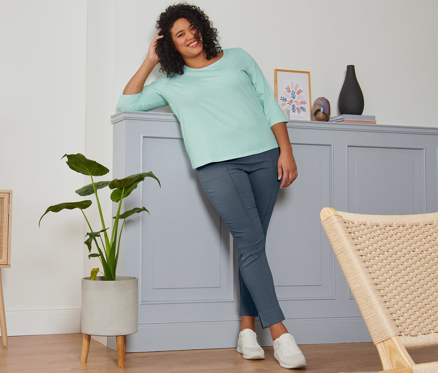 Une femme souriante, appuyée contre un mur, porte un t-shirt manches 3/4 menthe, un pantalon stretch bleu acier et des baskets en cuir velours.