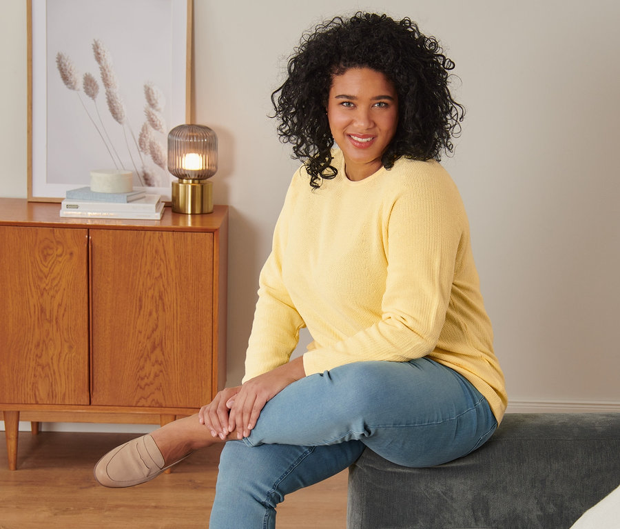 Femme assise sur un canapé gris portant un pull en maille jaune, un tregging et des loafers en cuir velours.