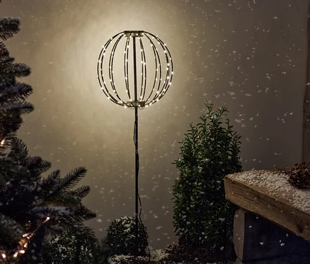 Une pique de jardin LED « Boule » illuminée se dresse entre des buissons enneigés.