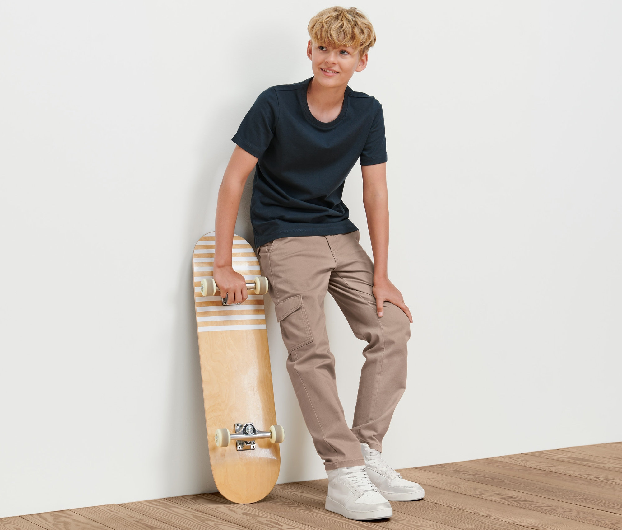Un garçon appuyé contre un mur tenant un skateboard. Il porte un pantalon cargo pour enfant.