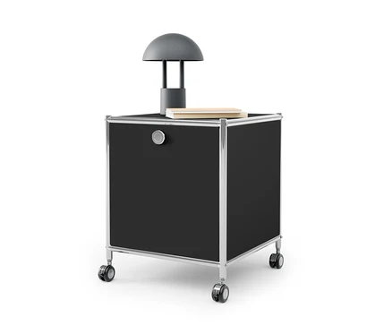 Table d’appoint noire avec tiroir en métal « CN3 » avec cadre argenté et roulettes. Une lampe grise et un livre sont posés sur la table.