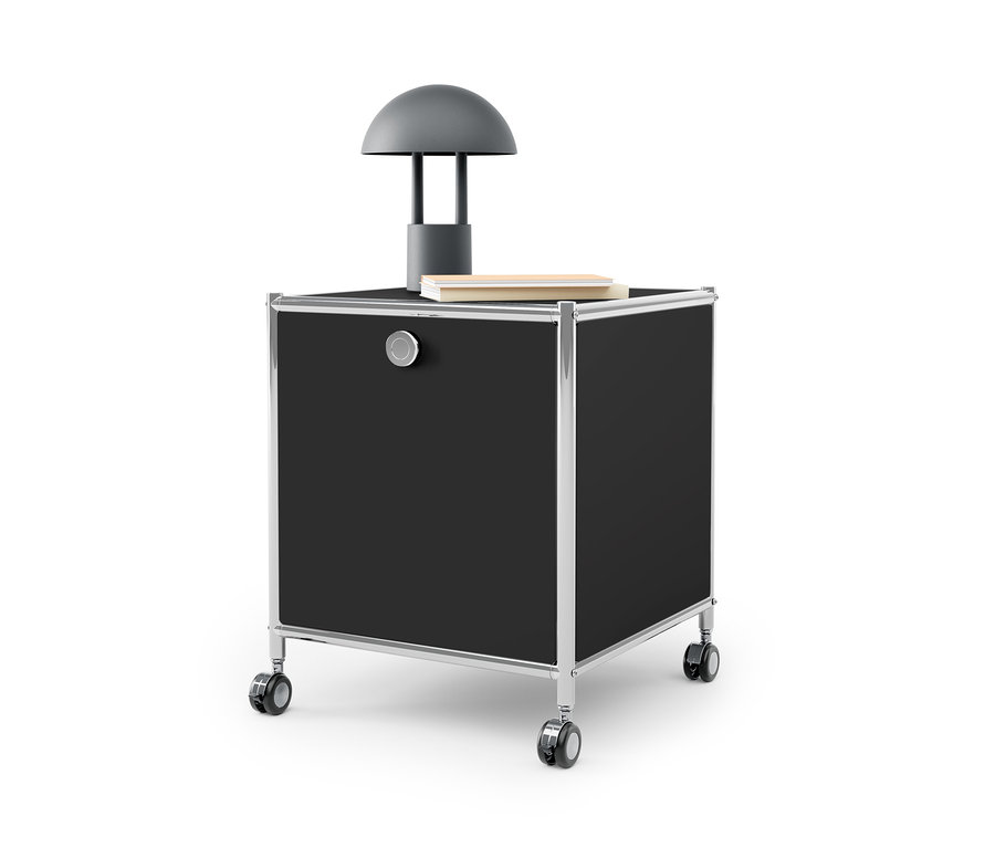 Table d’appoint noire avec tiroir en métal « CN3 » avec cadre argenté et roulettes. Une lampe grise et un livre sont posés sur la table.