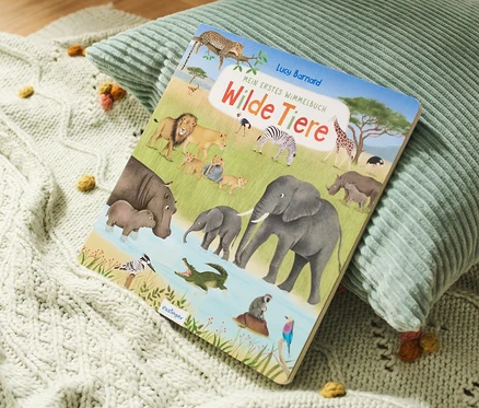 Le livre « Mein erstes Wimmelbuch – Wilde Tiere » est posé sur un coussin vert et une couverture tricotée.