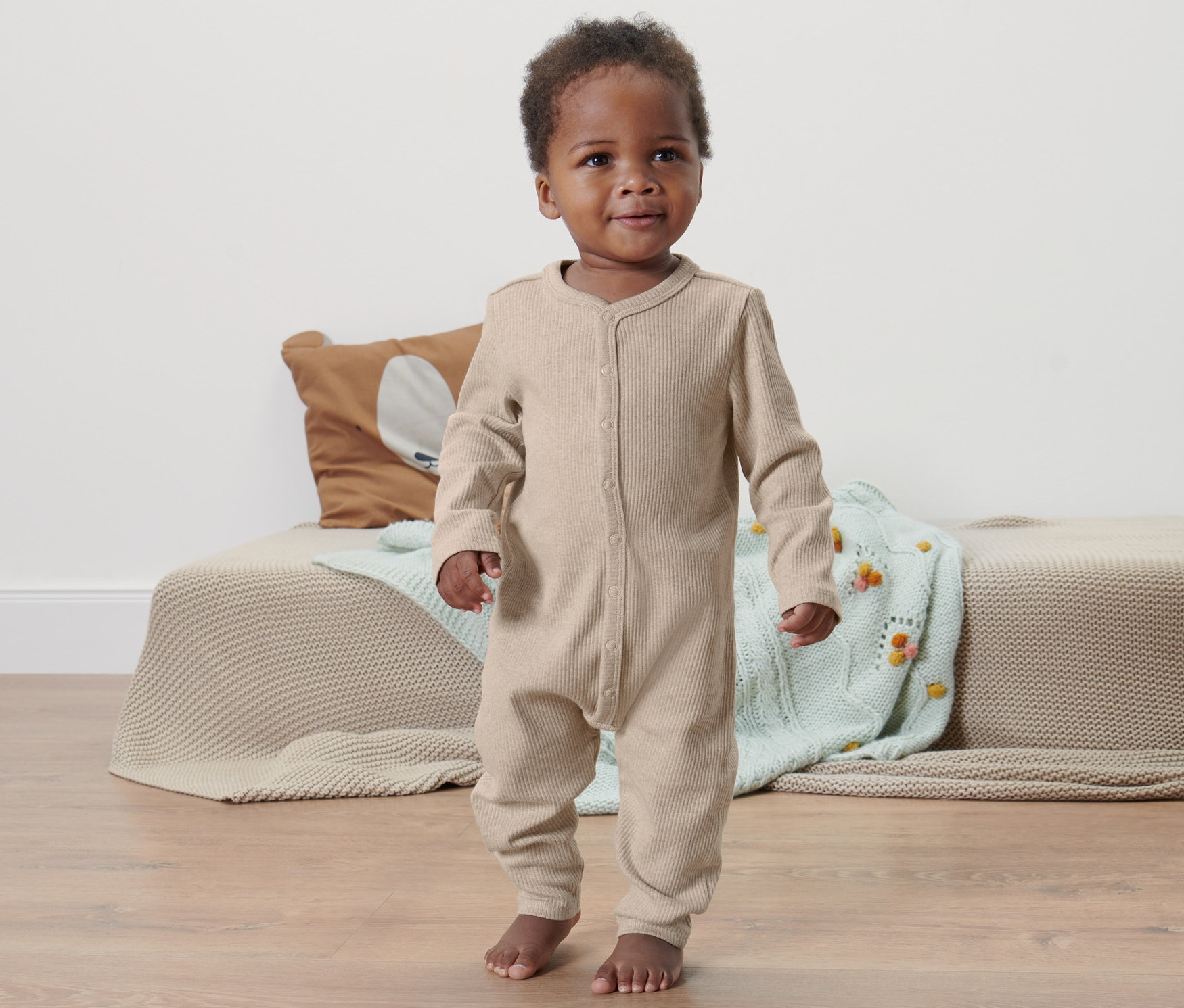 Un bébé à la peau foncée se tient sur le sol et porte une barboteuse.