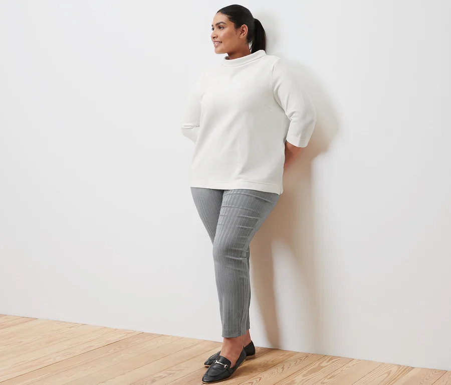 Une femme appuyée contre un mur porte un sweat-shirt manches 3/4 crème, un pantalon stretch et des loafers en cuir.