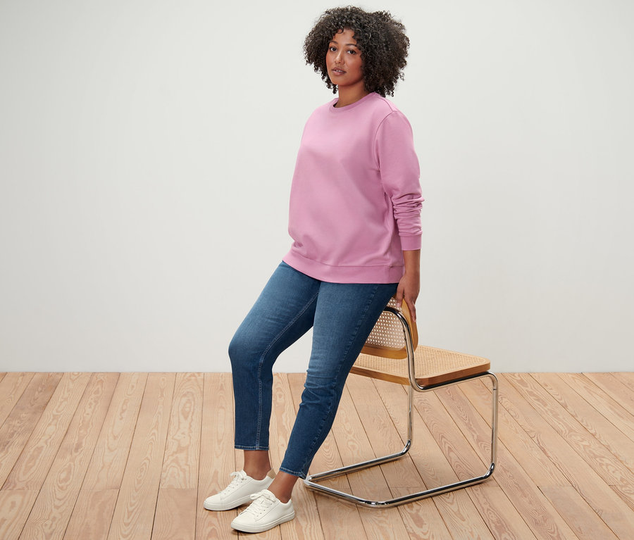 Une femme est assise appuyée contre une chaise, portant un sweat-shirt rose, un tregging « Fit Nelly » et des baskets blanches.