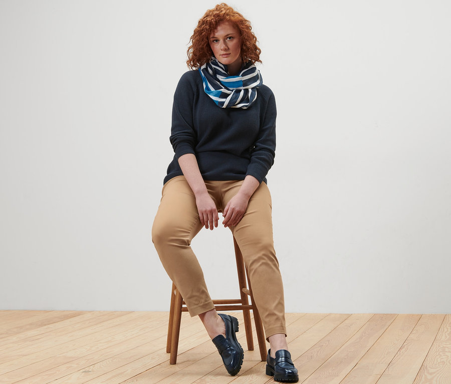 Une femme est assise sur un tabouret et porte un pull en maille bleu foncé, un col écharpe à rayures et un pantalon stretch.