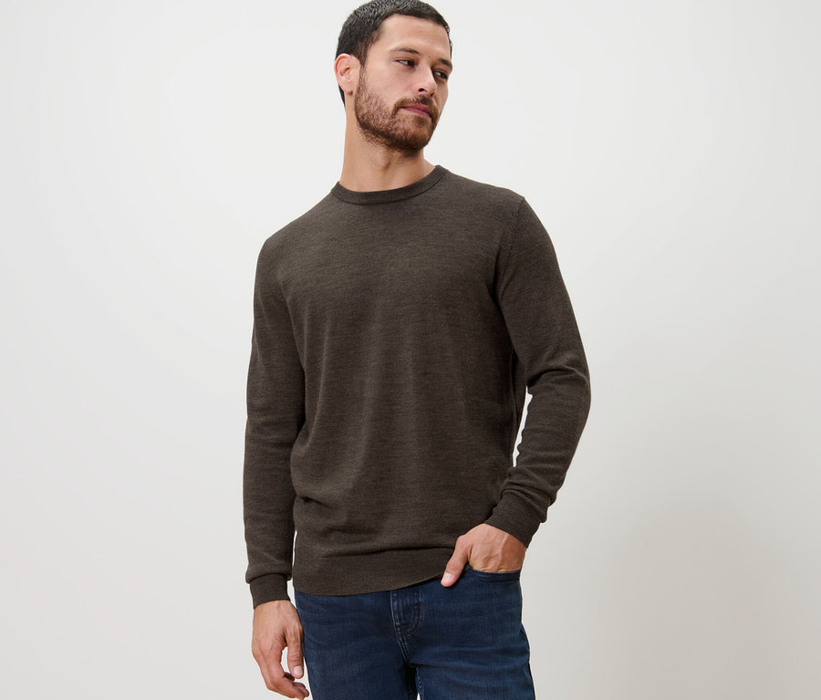 Un homme pose en pull en mérinos marron et en jean.