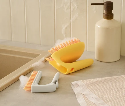 Une brosse à sol et carrelage 2 en 1 jaune et une brosse à détails blanche sont posées côte à côte sur un comptoir à côté d'un évier.