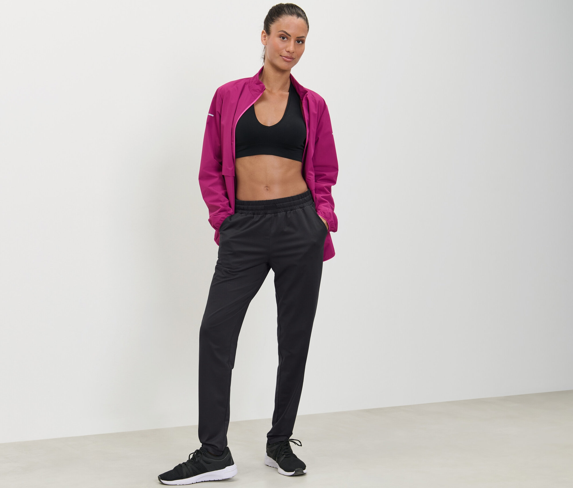 Une femme pose, les mains dans les poches d'un pantalon de sport noir.