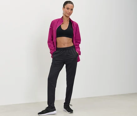 Une femme pose, les mains dans les poches d'un pantalon de sport noir.