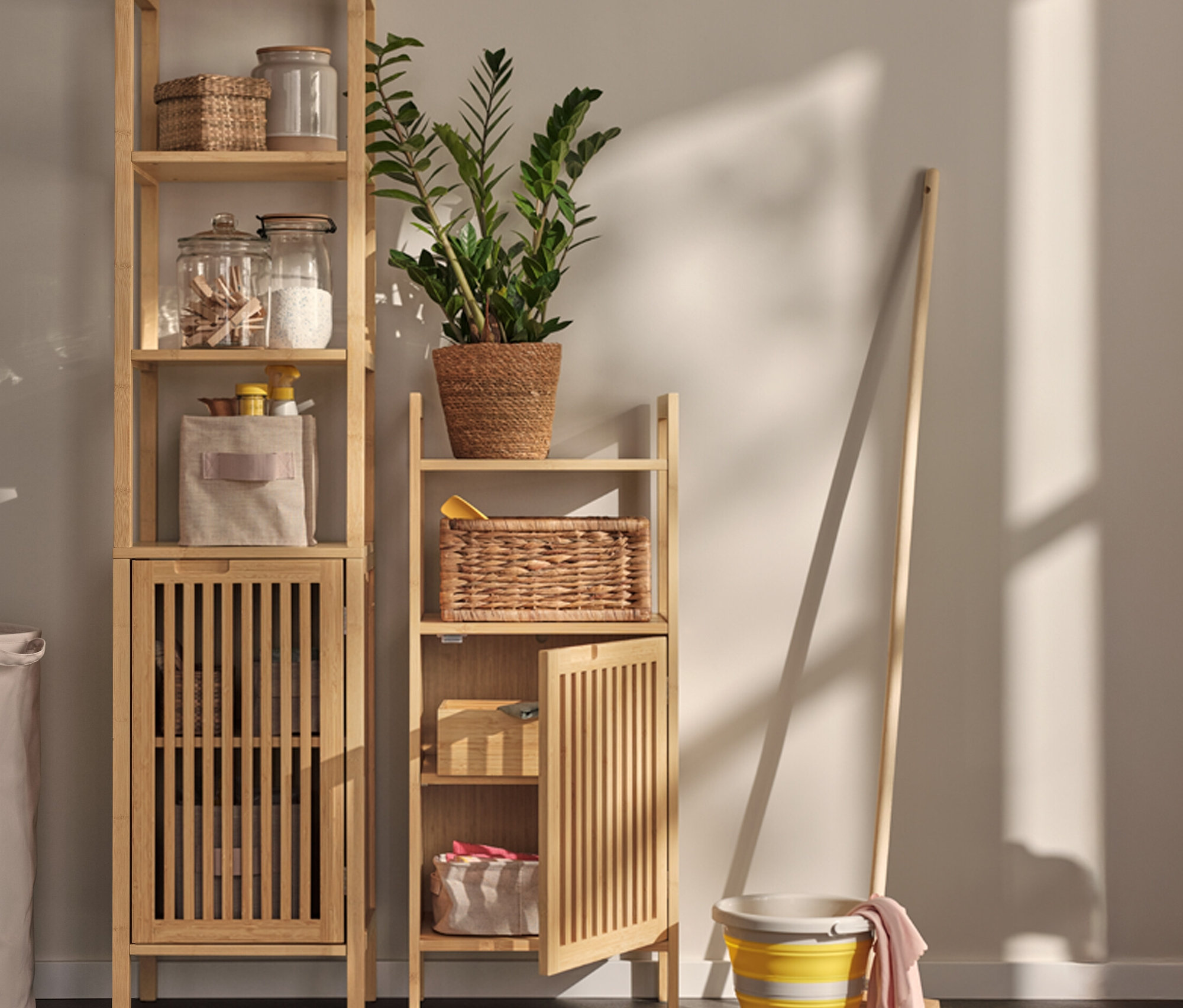 Deux étagères de rangement de style scandinave sont placées dans la pièce, remplies de bocaux de rangement, de paniers et de textiles. Un panier à linge et un seau de nettoyage avec une serpillière sont également visibles.