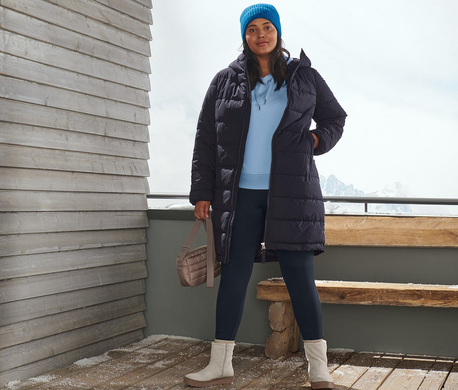 Une femme pose à l'extérieur sur une terrasse en bois avec un manteau matelassé bleu foncé, un bonnet en maille bleu, un pull à capuche, un legging thermique bleu, des bottines en cuir velours et un sac matelassé à bandoulière.