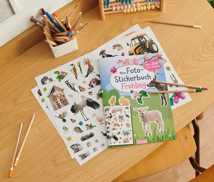 Sur une table sont posés un livre ouvert « Mein Foto-Stickerbuch – Frühling » (en allemand), une planche d'autocollants, des crayons de couleur et un boulier.