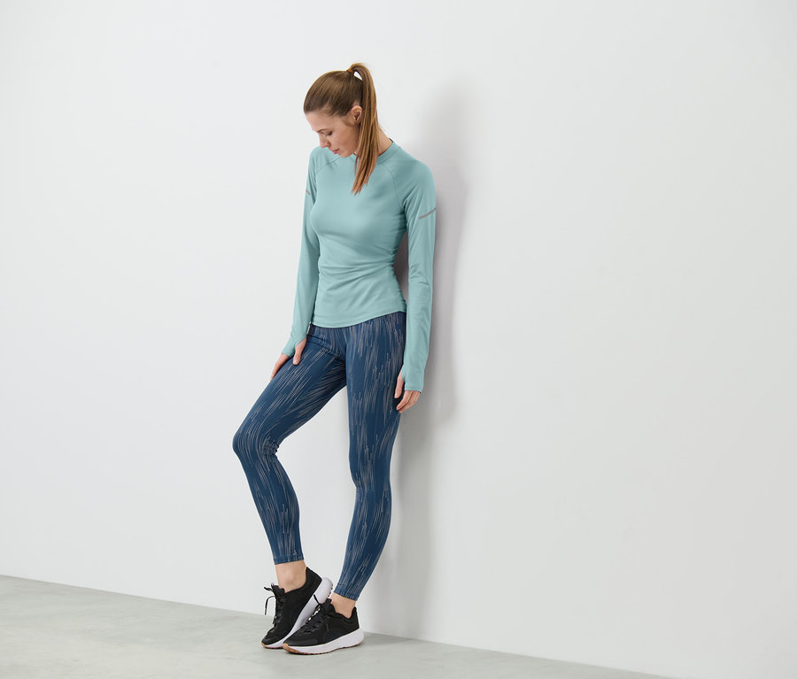 Une femme appuyée contre un mur porte un legging de sport réfléchissant argenté et un haut à manches longues.