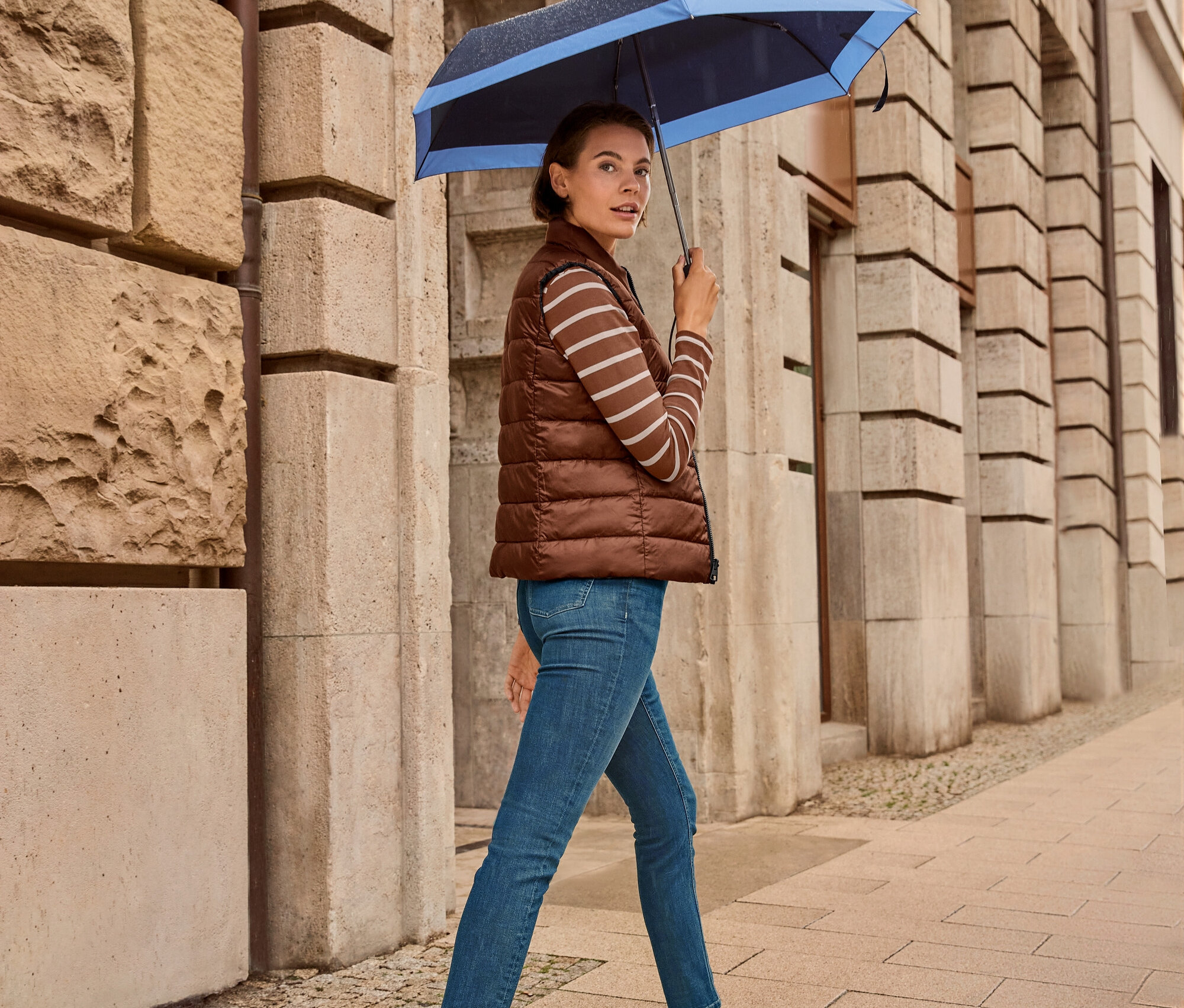 Une femme marche sur le trottoir avec un parapluie, elle porte un gilet matelassé réversible, un jean slim – Coupe « Emma », bleu foncé et des baskets en cuir.