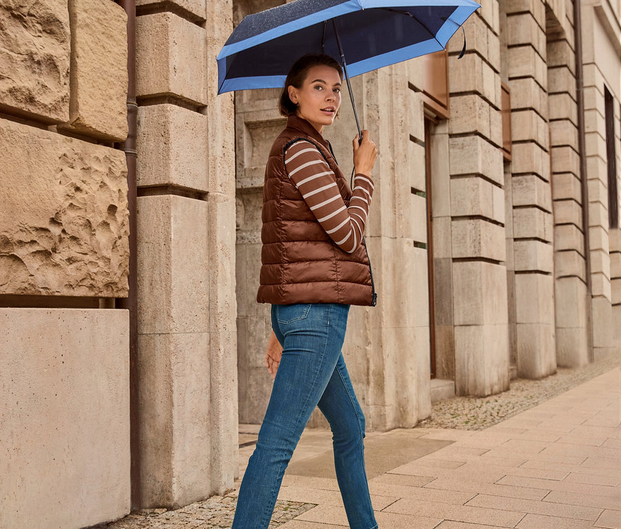 Une femme marche sur le trottoir avec un parapluie, elle porte un gilet matelassé réversible, un jean slim – Coupe « Emma », bleu foncé et des baskets en cuir.
