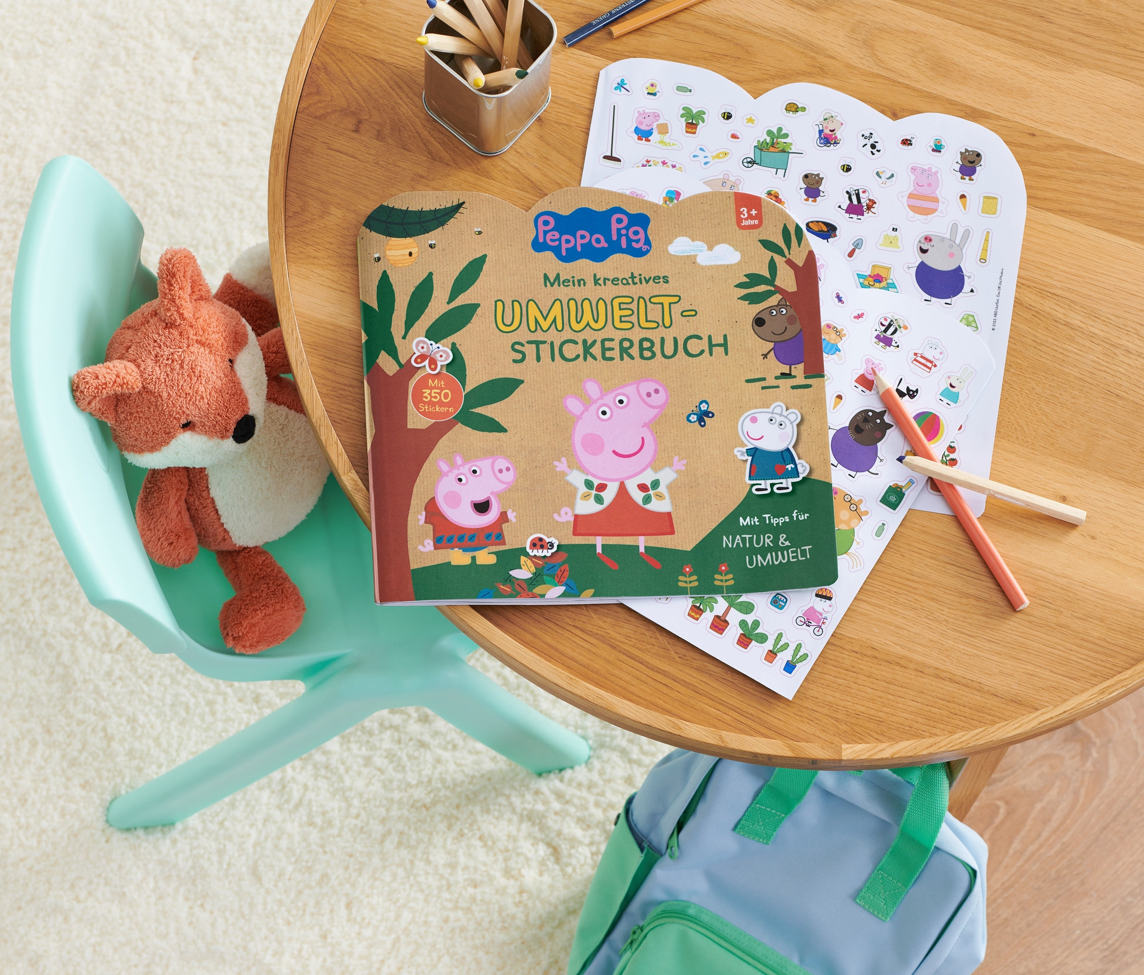 Sur une table sont posés un livre « Peppa Pig ‒ mein kreatives Umwelt-Stickerbuch » (en Allemand) et une feuille d'autocollants. Un renard en peluche est posé sur une chaise.