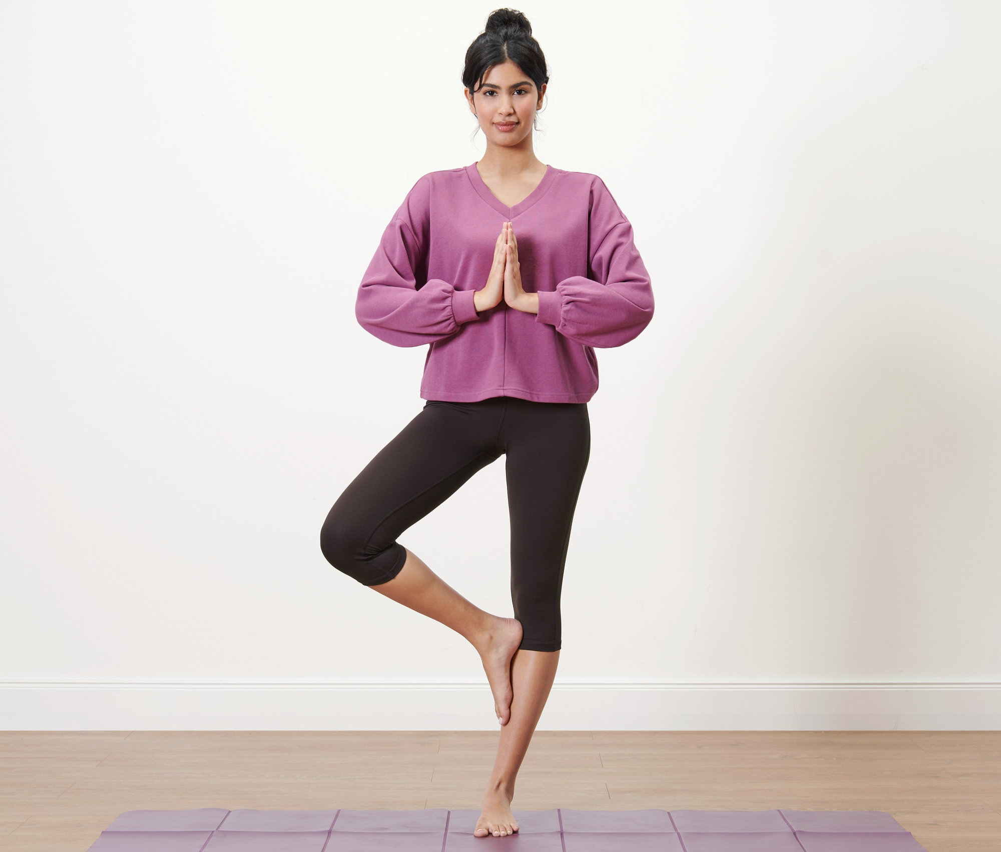 Femme debout dans la pose de l'arbre sur un tapis de yoga violet portant un collant de sport modelant 3/4.