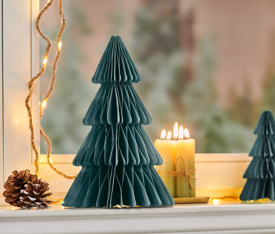 Un arbre en papier pliable vert foncé de taille moyenne est placé à côté d'une bougie allumée et d'une pomme de pin sur le rebord d'une fenêtre.