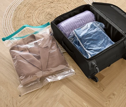 Un manteau beige dans un sachet de voyage sous vide, à côté d'une valise noire avec un pull violet et une veste en jean dans des sachets de voyage sous vide.