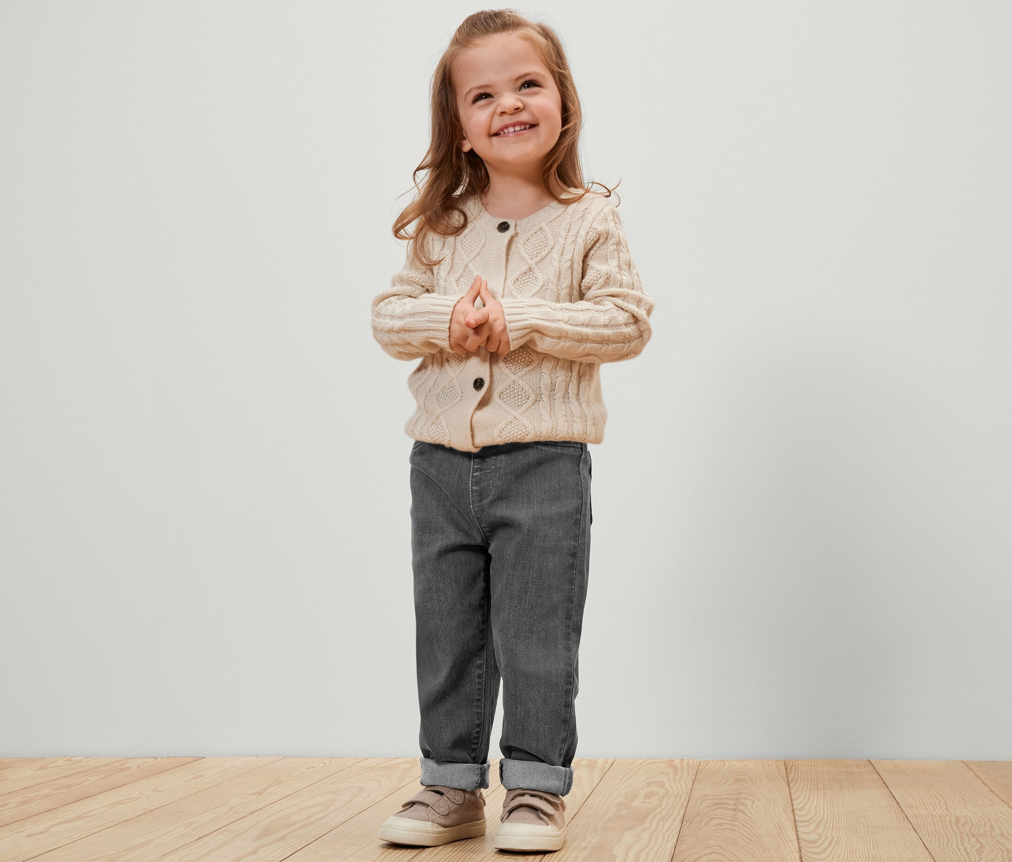 Petite fille souriante debout, les mains jointes. Elle porte un pantalon gris et un cardigan crème. Pantalon à enfiler pour enfant – Fit « Luca », gris.