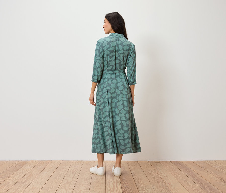 Une femme se tient dos à la caméra et porte une robe verte à motifs et des baskets blanches.