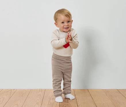 Un bébé aux cheveux blonds applaudit sur un plancher en bois devant un mur blanc.